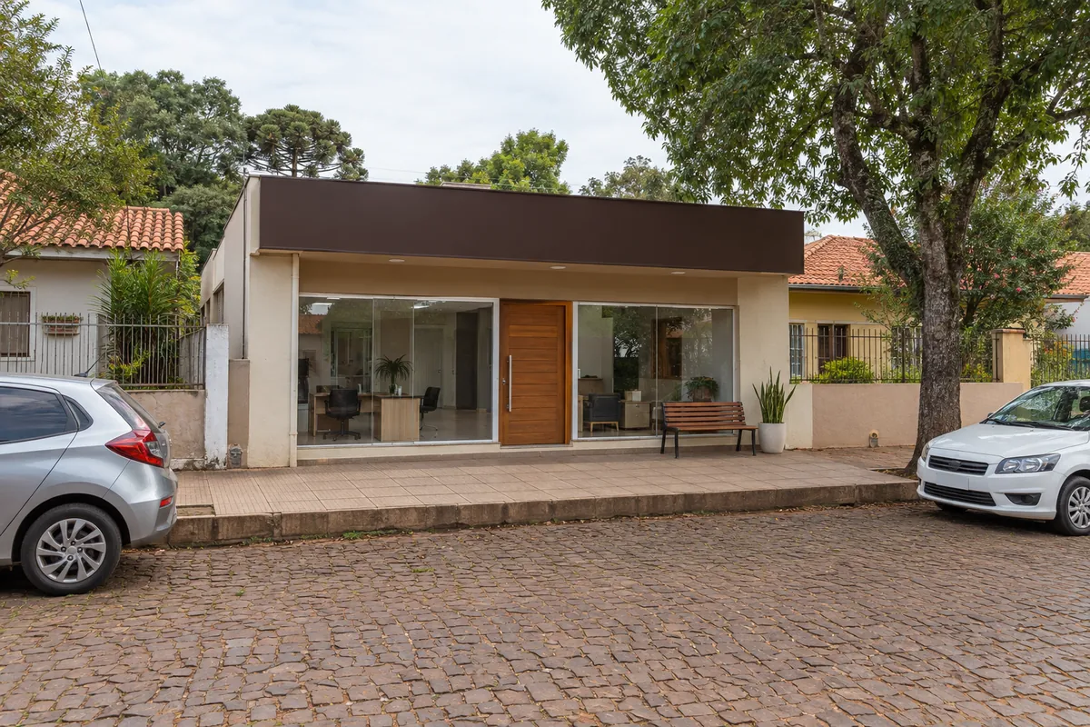 Fachada do escritório da imobiliária, com vitrine de vidro, porta de madeira, um banco e um vaso de planta, numa rua comercial calçada.